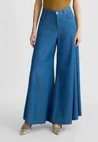 Weite blaue Denim-Hosen mit hohem Bund, die Taschen und einen vorderen Knopfverschluss aufweisen. Der Stoff hat eine glatte, einheitliche Textur.