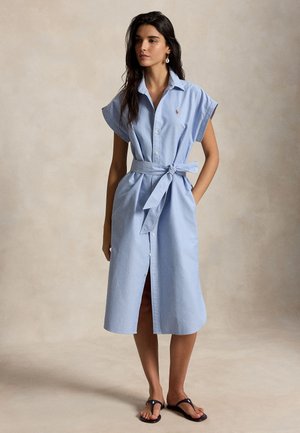 Kobieta ubrana w jasnoniebieską sukienkę koszulową z paskiem i krótkimi rękawami oraz czarne sandały, stojąca na tle jednolitego beżowego tła.