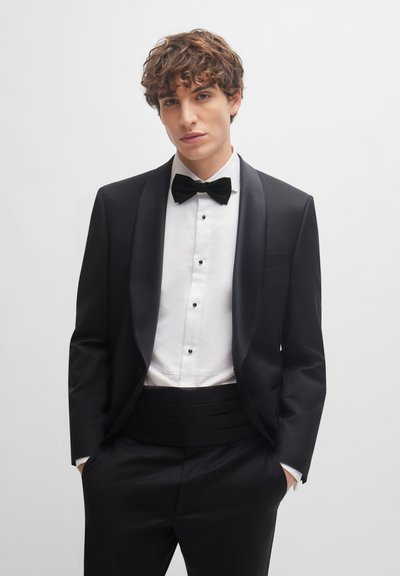 Veste de costume noire avec revers en satin, chemise blanche à boutons, nœud papillon noir et pantalons noirs avec ceinture assortie. Texture lisse sur l'ensemble.