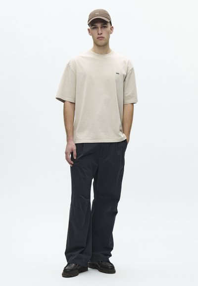 Jeune homme portant une casquette beige et un T-shirt beige surdimensionné avec un pantalon noir et des chaussures noires, debout contre un fond clair uni.