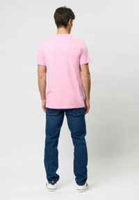 Różowy bawełniany t-shirt z krótkimi rękawami, o prostym kroju i minimalną ilością szwów; noszony z niebieskimi dżinsami i białymi trampkami.