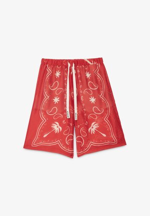 Mujer con el pelo corto y oscuro que lleva una chaqueta marrón, falda roja con estampado, bufanda roja y mocasines blancos, de perfil con las manos en los bolsillos.