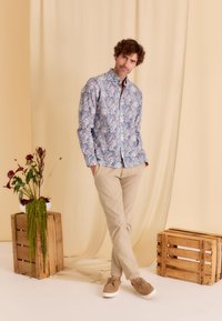 Lichtblauw bloemenoverhemd met roze accenten, beige broek en bruin schoeisel, tegen een zachte gele achtergrond met houten kisten.