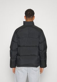 Doudoune noire avec un col montant et un design matelassé, présentant des panneaux horizontaux contrastants, portée avec un jean bleu clair.