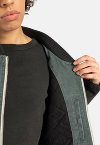 Groene suède jas met een gewatteerde zwarte voering, een geribde zwarte kraag en een zilveren rits, gedragen over een zwarte geribde lange mouwen shirt.