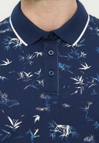 Μπλε ναυτικό πόλο μπλουζάκι με λευκά και μπλε αφηρημένα φύλλα, διαθέτει ριμπ ύφασμα στον γιακά με λευκό περίγραμμα και τρία μπλε κουμπιά.