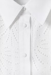 Col de chemise blanc avec boutons recouverts de tissu et motifs floraux brodés complexes le long de la patte de boutonnage avant.