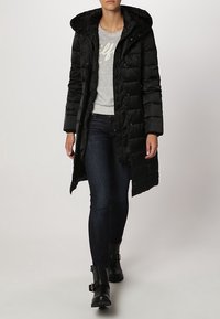 Schwarzer Puffer-Mantel mit Kapuze, gestepptes Design, lange Länge, mit Druckknöpfen versehen. Getragen über einem grauen Pullover und dunklen Jeans.