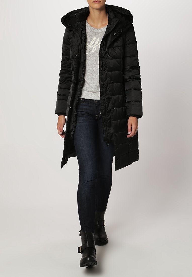 Schwarzer Puffer-Mantel mit Kapuze, gestepptes Design, lange Länge, mit Druckknöpfen versehen. Getragen über einem grauen Pullover und dunklen Jeans.