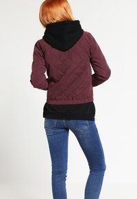 Giacca invernale bordeaux trapuntata con cappuccio nero, vita aderente e finitura testurizzata, abbinata a jeans skinny blu.