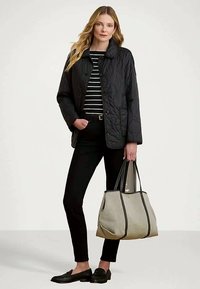 Γυναίκα φορώντας μαύρο καπιτονέ μπουφάν, μαύρο και λευκό ριγέ μπλουζάκι, μαύρο παντελόνι, μαύρες loafer, κρατώντας μεγάλη μπεζ τσάντα ώμου με μαύρους ιμάντες.