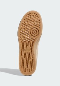 Adidasova podplatka športnih copat z gumijasto oblogo, ki vključuje teksturiran vzorec za oprijem in osrednji krožni emblem. Svetlo rjava barva.