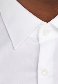 Camisa blanca con cuello, de textura suave, con cierre de botones y diseño clásico. Las puntas del cuello son angulares y bien definidas.