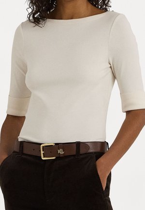 Mujer que lleva una camiseta ajustada color crema con mangas hasta el codo, pantalones marrón oscuro y un cinturón de cuero marrón con una hebilla dorada.