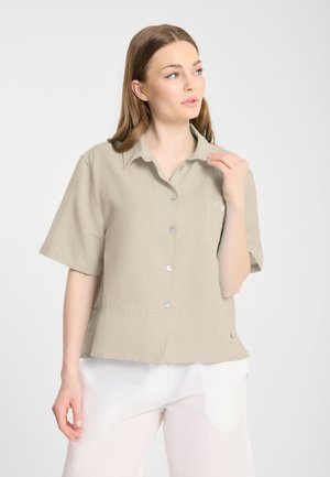 Kobieta o jasnobrązowych włosach, ubrana w beżową koszulę na krótki rękaw z guzikami i białe spodnie, patrząca w bok.