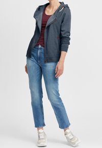 Sudadera con capucha en gris oscuro con cremallera, cuenta con cordones blancos y acentos marrones; combinada con jeans rectos de color azul claro y zapatillas beiges.