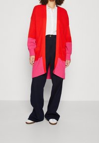 Kobieta ubrana w czerwono-różowy dziergany sweter, białą koszulę na guziki, czarne spodnie z szerokimi nogawkami oraz białe buty na jednolitym tle.