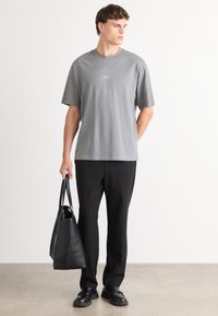 Grå kortermet T-skjorte med rund hals, som har en liten logo på brystet. Brukt med svarte bukser og holder en svart totebag.