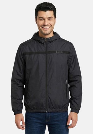 Homme souriant portant une veste à capuche noire zippée avec des poignets élastiques et un jean bleu sur un fond uni.