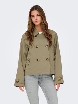 Woman with long brown hair wearing an oversized olive green double-breasted jacket and light grey jeans against a plain background.