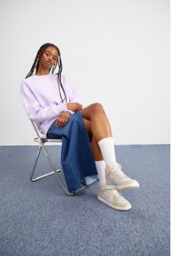 Ljuslila sweatshirt med Nike-logga, denimkjol, vita strumpor och beige sneakers med ljusblå och rosa accenter, sittande på en silverstol.