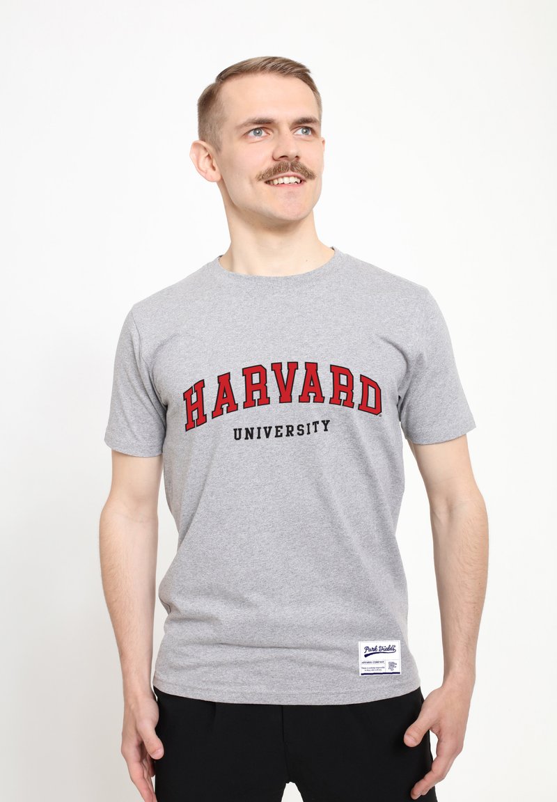 Šedé bavlněné tričko s nápisem "HARVARD" v červeném písmu a "UNIVERSITY" v černém písmu níže. Krátké rukávy s štítkem na dolním lemu.