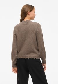 Pruun kudumissügisese särgiga, millel on pikad varrukad, laineline kaelus ja äär, ning millel on tekstuurne muster mansettidel ja alumisel äärel.