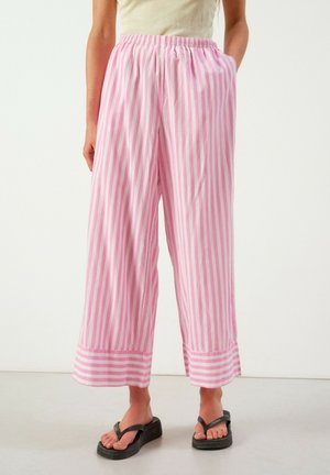 Vrouw die losse roze-wit verticale gestreepte wijde broek met omgeslagen zoom en zwarte plateauflipflops draagt, staand op een effen vloer.