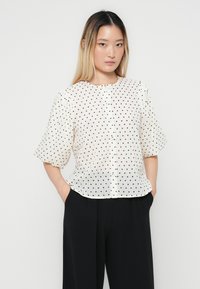 Blouse blanche à pois noirs, manches courtes bouffantes, col rond, fermeture à boutons sur le devant, et tissu léger. Associée à un pantalon noir.