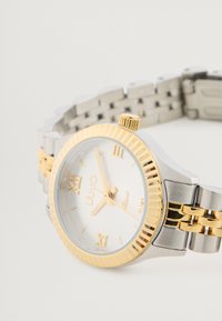 Ezüst és arany színű fém karóra fehér számlappal, római számokkal jelölt órabeoszt�ással és bordázott arany kerettel. Különböző fémből készült karkötővel rendelkezik.