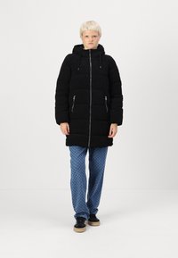 Manteau matelassé noir avec une capuche, poches zippées et une fermeture éclair à l'avant. Assorti avec un pantalon bleu à motifs et des baskets noires à semelle en gomme.