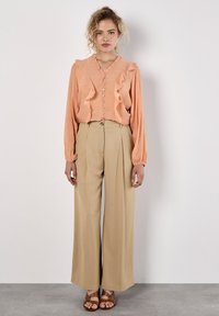 Blouse en pêche avec volants et boutons sur le devant, associée à un pantalon large taille haute beige et des sandales à lanières marron. Tissu texturé sur l'ensemble.