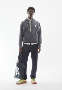 Grijze hoodie met wit logo, beige trekkoorden, donkere broek, kleurrijke hybride sneakers en een groene tote bag met een grote witte letter "A."