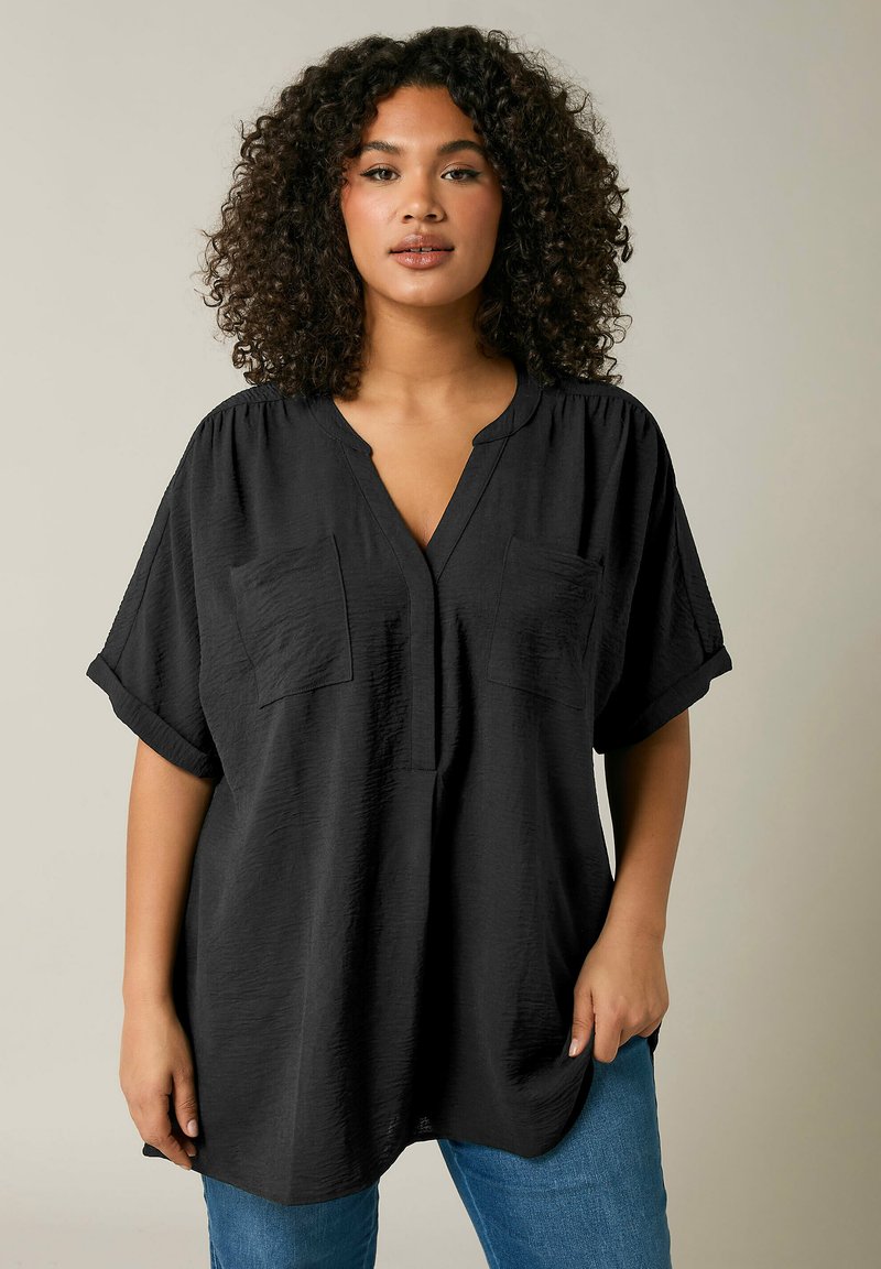 Blusa nera a maniche corte con scollo a V, due tasche sul petto e un tessuto testurizzato. Abbinata a jeans blu.