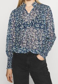 Donna che indossa una blusa trasparente blu navy con un motivo floreale pastello e spalle arricciate, abbinata a jeans neri a vita alta, con una mano nella tasca.