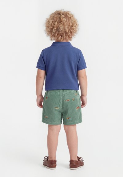 Παιδί με ξανθά σγουρά μαλλιά που φορά μπλε πόλο μπλουζάκι, πράσινα κεντημένα σορτς και καφέ παπούτσια, στέκεται με την πλάτη προς τα εμπρός σε λευκό φόντο.