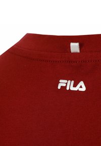 Camiseta de algodón roja con cuello redondo; presenta un logo bordado en blanco en la parte superior izquierda. Textura suave y diseño sencillo.