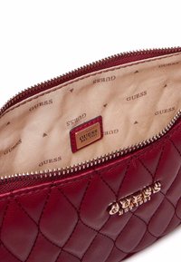 Pochette sintetica trapuntata color bordeaux con logo "GUESS" in tono dorato, chiusura con zip e interno beige con un motivo ripetuto "GUESS".