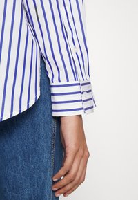 Hvit skjorte med blå striper, knappet mansjett, glatt tekstur, kombinert med denim. Mansjetten har et mønster med vekslende striper og knappedetalj.