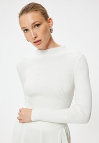 Top à manches longues blanc côtelé avec un col haut. Présente une silhouette ajustée et une texture douce, associé à une jupe blanche lisse.