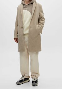 Abrigo de lana beige con dos botones, superpuesto a una sudadera de color claro con un patrón curvado, combinado con pantalones claros y zapatillas negras.