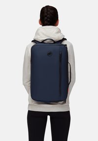 Marinblå ryggsäck med rektangulär form, tillverkad av slät material, med en framfick med dragkedja och en logodetalj.