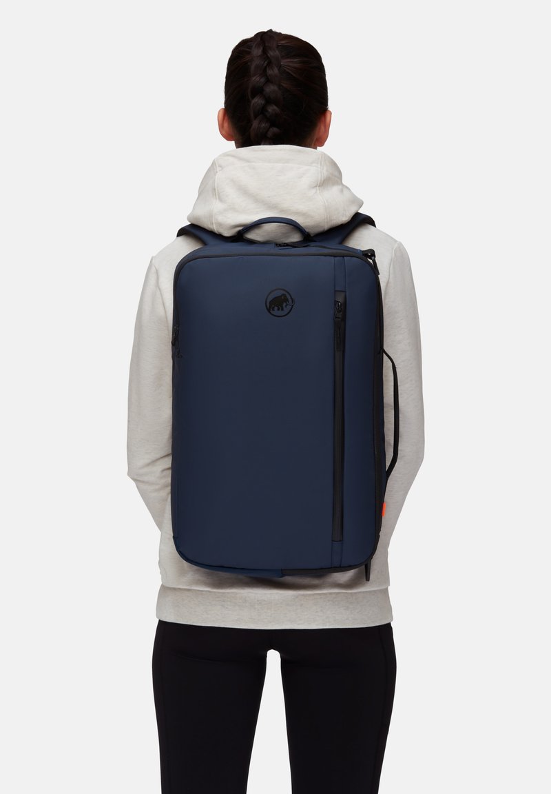 Marinblå ryggsäck med rektangulär form, tillverkad av slät material, med en framfick med dragkedja och en logodetalj.