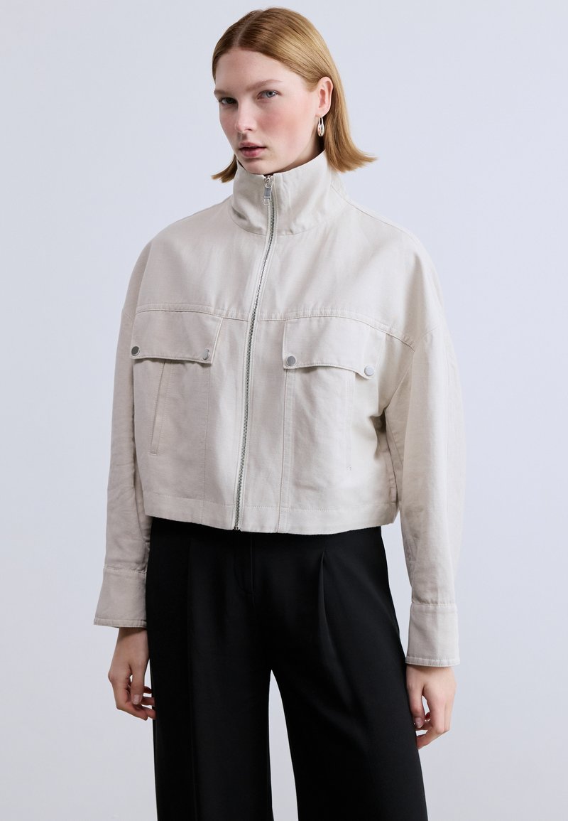 Κοντό, ανοιχτό μπεζ σακάκι με ψηλό γιακά, φερμουάρ μπροστά και δύο τσέπες στο στήθος. Κατασκευασμένο από υφασμα με υφή και ελάχιστες ραφές.