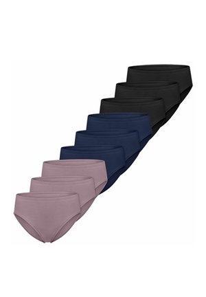 Frau mit lockigem, dunklem Haar, die einen marineblauen BH und passende Unterwäsche trägt, vor einem schlichten hellen Hintergrund steht und seitlich schaut.