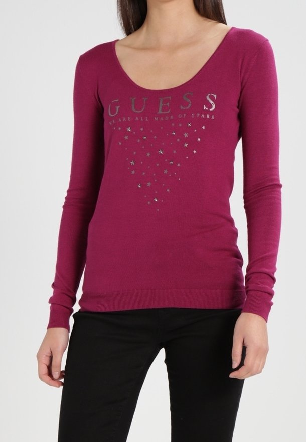 Femme portant un haut moulant à manches longues magenta avec des motifs "GUESS" et étoiles, associé à un pantalon noir sur un fond uni.
