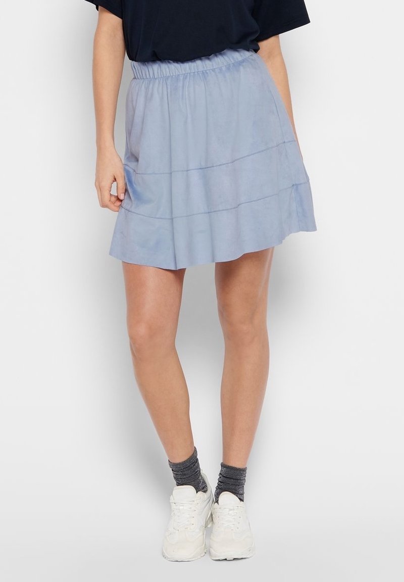 Femme portant une mini-jupe bleu clair à taille élastique, des chaussettes gris foncé, des baskets blanches et un haut noir, montrée de la taille aux pieds.