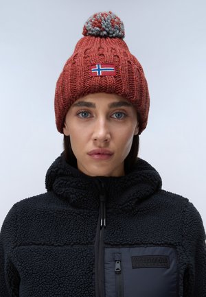Berretto lavorato a maglia rosso con un pom-pom grigio e una toppa con la bandiera norvegese. Indossato con una giacca in pile nera con cappuccio e cerniera.