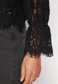 Black crocheted lace top with textured floral patterns, bell sleeves, and fringe detailing at the hem, worn over gray denim.
