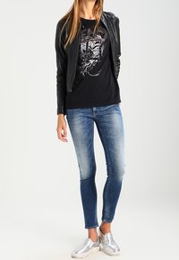 Žena oblečená v černé kožené bundě, černém tričku s potiskem, vybledlých skinny džínách a stříbrných slip-on botách, stojící na bílém pozadí.